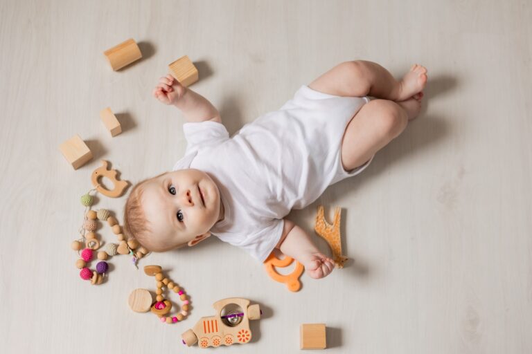 cute baby in white bodysuit lies on his back on the floor among rattles and wooden educational toys