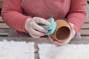 Hands of senior woman moisturizing handmade clay vase by sponge for further coloring with glaze